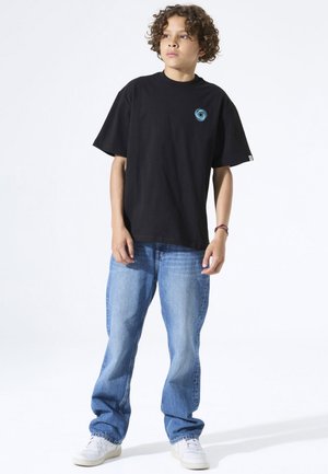 Black cotton t-shirt with short sleeves, featuring a blue circular logo on the chest, paired with light blue denim jeans and white sneakers.
