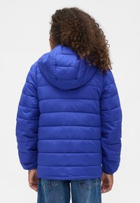 Blå vadderad jacka med huva, med quiltad textur och elastiska muddar, kombinerad med blå denimjeans. Bilden visar jackans baksida med fokus på designen.