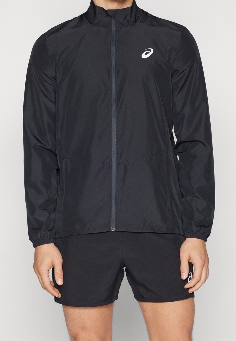 Homme portant une veste de sport noire à fermeture éclair et un short de sport noir, tous deux avec des logos Asics blancs, se tenant devant un fond uni.