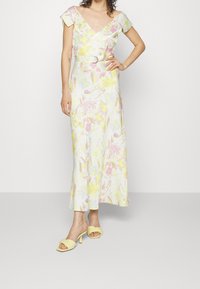 Bloemenmidi-jurk in wit, met pastelroze, gele en blauwe patronen, kapmouwen en een tailleceintuur. Gecombineerd met gele sandalen met hak.