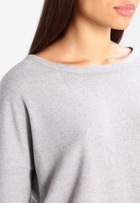 Jasnoszary sweter o szerokim dekolcie, miękkiej fakturze i luźnym kroju. Materiał wygląda na dziany z lekko widocznymi włóknami.