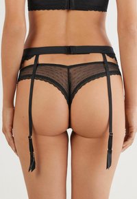 Schwarzer Spitzenstring mit gepunktetem Mesh, ausgestattet mit gewelltem Saum, verstellbaren Trägern und Schleifenakzenten auf der Rückseite.