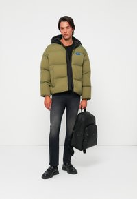 Olive green puffer jacket with black accents, front zip, and hood; paired with dark jeans and a black backpack. Smooth texture and boxy shape.