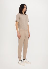 Beige strikket genser med kort erme, mønstrete tekstur og kontrastfarge i brunt kantbånd, sammen med beige joggebukser og hvite sneakers med svarte detaljer.