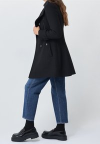 Cappotto in misto lana nero con vestibilità su misura, ampio colletto, bottoni doppi, tasche laterali e una texture liscia, abbinato a jeans blu e scarpe nere.