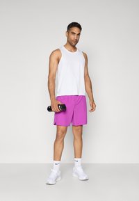 Homme portant un haut de sport sans manches blanc et un short violet, tenant une bouteille d'eau noire, se tenant devant un fond uni.