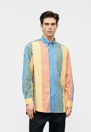 Man wearing long-sleeve button-up shirt with vertical stripes in blue, green, orange, yellow, and white, standing against a plain background.