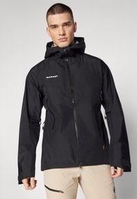 Veste imperméable noire avec capuche, fermeture éclair et deux poches latérales zippées. Présente un logo sur la poitrine et des poignets ajustables.
