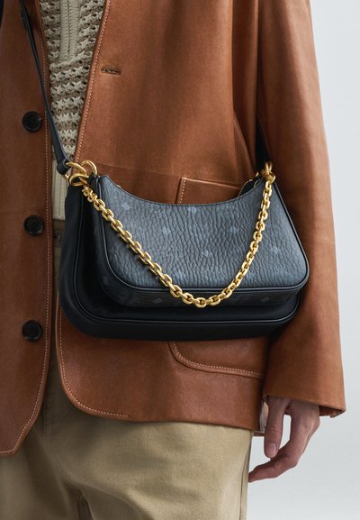 Osoba ve světlých kalhotách, krémovém pleteném svetru a hnědé kožené bundě drží dvě černé kožené kabelky; jedna má zlatý řetízek.