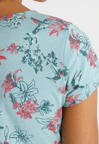 Camisa de color azul claro con estampado floral de flores rosas y verdes y colibríes; mangas cortas con escote redondeado.