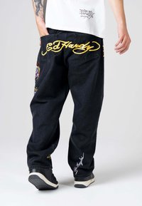 Jeans in denim nero con ricami gialli che riportano "Ed Hardy". Patches decorativi con disegni colorati sulle tasche posteriori. Vestibilità ampia.