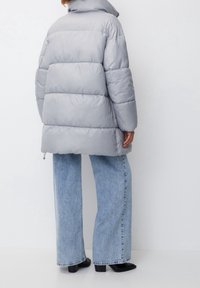 Chaqueta de plumas gris con cuello alto, corte oversize y diseño acolchado, combinada con unos vaqueros de pierna ancha en azul claro y zapatos negros.