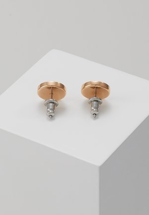 Paire de boucles d’oreilles puces rondes en métal or rose et argent placées sur un piédestal géométrique blanc sur un fond clair.