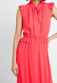 Korallfarbendes ärmelloses Kleid mit Rüschendetails an den Schultern, Schleifenkragen, geraffter Taille und fließendem Rock. Glattes Material mit einer weichen Textur.