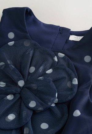 Marineblå kjole med polkaprikmønster, der har en stor mesh-blomsterdetalje og en glat stoftekstur. Mærkat med mærkenavn synlig.
