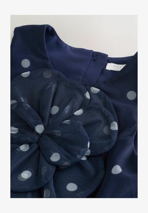 Vestido azul marino con patrón de lunares, que presenta un gran detalle de flor de malla y una textura de tela suave. Etiqueta visible con el nombre de la marca.