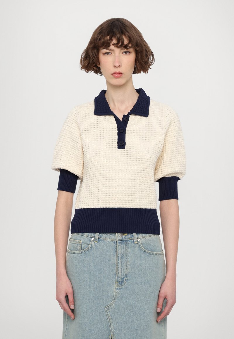 Pull tricot crème et marine avec col polo, manches bouffantes courtes et devant boutonné. Texture gaufrée sur le corps, ourlet et poignets côtelés.