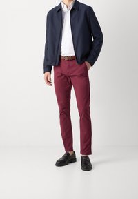 Giacca bombardiere navy, camicia bianca, pantaloni chinos bordeaux e mocassini neri con dettagli in oro. Tessuto liscio, design aderente e tonalità a contrasto.