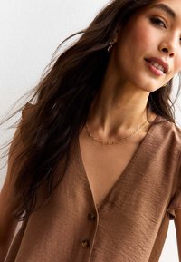 Blouse marron à col en V avec des manches courtes et deux boutons à l'avant. Tissu texturé avec un subtil éclat, associé à un collier en chaîne doré.