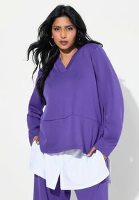 Pullover viola a scollo a V con maniche lunghe, realizzato in tessuto morbido. Indossato sopra una camicia bianca, presenta un design a strati e una vestibilità comoda.