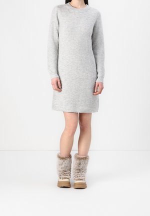 Kobieta ubrana w jasnoszarą, długą dzianinową sukienkę z długim rękawem i beżowe zimowe buty ocieplane futrem, stojąca na białym tle.