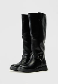 Bottes en cuir noires à hauteur de genou avec une finition lisse, arborant une sangle latérale et un détail de boucle, ainsi qu'une semelle plate pour plus de stabilité.