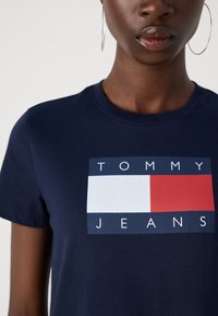 Tmavej pleti osoba s veľkými kruhovými náušnicami a námornícky modrým tričkom s hranatým logom Tommy Jeans v bielej, červenej a modrej farbe.