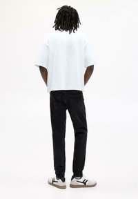 Person med dreadlocks står vendt væk, iført en hvid oversized t-shirt, sorte jeans og hvide sneakers med sorte accenter.