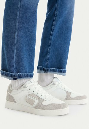 Białe i beżowe sneakersy noszone z białymi skarpetkami i podwiniętymi niebieskimi dżinsami na jednolitym jasnym tle.