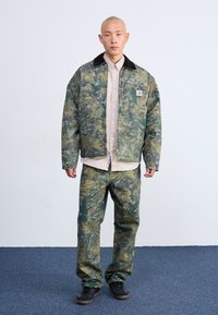 Chaqueta con patrón de camuflaje y cuello negro, combinada con pantalones a juego. Ambos están hechos de tela texturizada. Camiseta beige lisa debajo.
