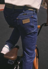 Jeans din denim albastru cu o etichetă cu logo-ul Wrangler, cu croială slim, cusături contrastante și nituri din cupru, asortate cu un șa de bicicletă maro.