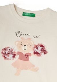 Světle béžové tričko s potiskem kreslené medvědice v růžové sukni s pompony, doplněné o růžové a burgundské odstíny.