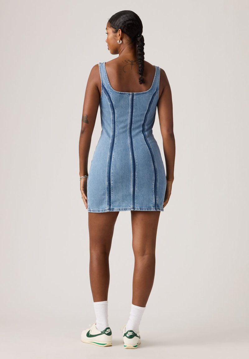 Denim minikjole med tætsiddende design, blå farve og lodrette sømdetaljer. Båret med hvide sokker og sneakers med grønne accenter.