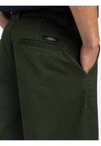 Grüne Baumwollmischungshose mit glatter Textur, ausgestattet mit Seitentaschen und einem schwarzen Markenlabel auf der Gesäßtasche.