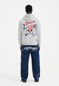 Gray hoodie featuring a large skull with a heart and banners reading "LOVE KILLS SLOWLY" on the back. Paired with blue jeans and black shoes.