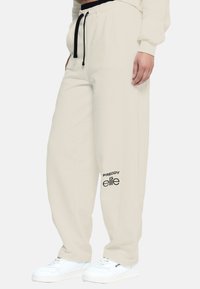 Pantaloni sportivi beige con cordino nero e logo stampato. Realizzati in materiale morbido, presentano una vestibilità rilassata e orli a cucitura. Indossati con sneakers bianche.