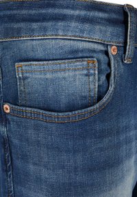 Tummansinisen denimkangasta tehdyt farkut, joissa on pieni takatasku, kuparinvärisiä niittiyksityiskohtia, näkyvää ommelta ja hieman kulunut rakenne.