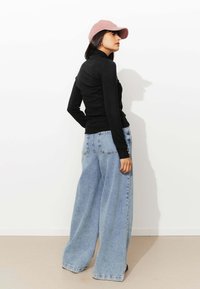 Un pull noir ajusté à col roulé et un pantalon en denim oversize bleu clair. Le modèle porte une casquette rose. Salle avec des murs blancs unis et un sol beige.