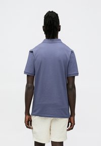 Ühevärviline sinine polosärk, millel on voltidega krae ja lühikesed varrukad, kombineeritud heledates toonides tekstureeritud lühikestega. Vaade tagantpoolt.