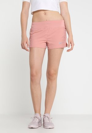 Frau trägt hellrosa Sportshorts mit kleinem grauem Nike-Logo und blassrosa Laufschuhen, steht vor einem einfarbigen Hintergrund.
