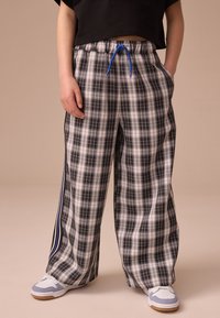 Pantalon large à carreaux noirs et blancs avec une ceinture élastique et un cordon de serrage bleu. Des rayures latérales apportent des détails. Porté avec des baskets blanches et bleues.