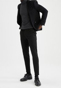 Musta bleiseri harmaan pooloneuleen päällä, yhdistetty mustiin slim fit -housuihin ja mustiin nauhallisiin kenkiin. Sileä kangas ja räätälöity istuvuus.