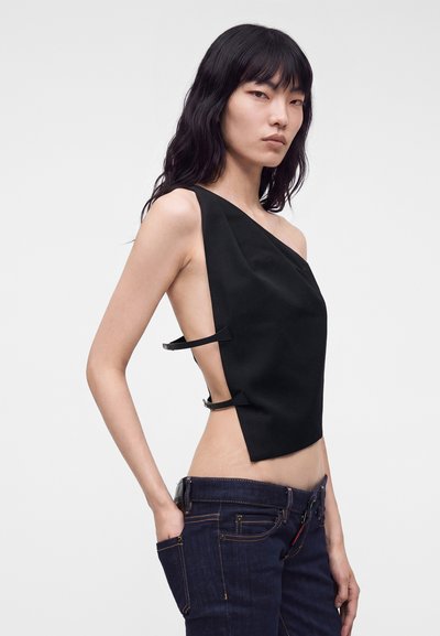 Jeune femme aux longs cheveux noirs portant un haut noir asymétrique sans manches avec des bretelles sur les côtés et un jean bleu foncé, la main dans la poche.