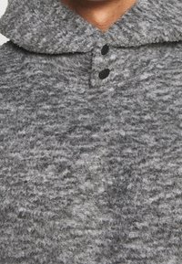 Maglione in pile grigio testurizzato con colletto, dotato di due bottoni neri per la chiusura. Il materiale appare morbido e con un motivo leggermente irregolare.