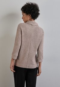 Maglione beige a maglia con colletto alto e maniche a tre quarti, caratterizzato da un motivo testurizzato e un orlo leggermente curvato sul retro.