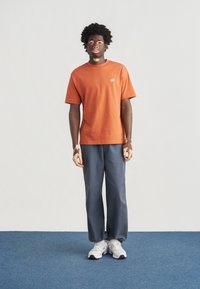 Πορτοκαλί βαμβακερό μπλουζάκι με κοντά μανίκια, λογότυπο στο στήθος. Γκρι παντελόνια με φαρδιά μπατζάκια και λευκά αθλητικά παπούτσια με μαύρες λεπτομέρειες. Στέκεται πάνω σε μπλε χαλί.