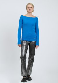 Blauwe, off-the-shoulder trui gemaakt van zachte stof, gecombineerd met glanzende, zilveren uitlopende broek. De schoenen hebben een chunky zool met kettingaccenten.