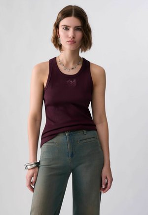 Jeune femme aux cheveux courts bruns portant un haut sans manches violet foncé, un pantalon vert, un collier en argent et des bracelets en argent, debout contre un mur blanc.