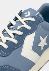 Blue sneaker featuring a textured suede and fabric upper, white star logo, and cream laces. It has a curved seam design with contrast stitching.
