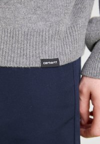Grey knit sweater with ribbed cuffs, featuring a black label with "carhartt." Paired with navy blue pants, showing smooth texture.